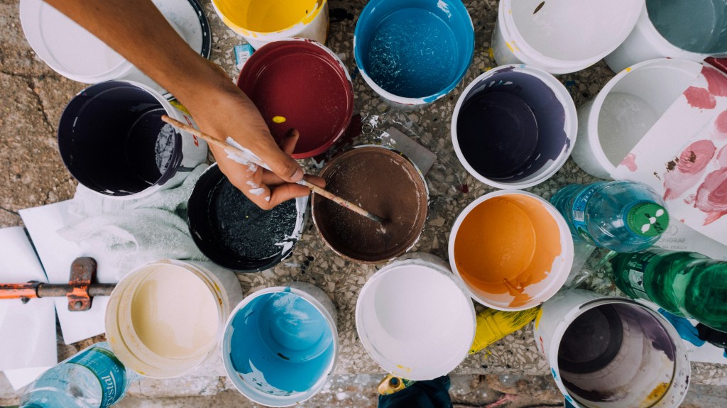 Collection of cups with various colors of paint with a person's hand holding a paint brush.